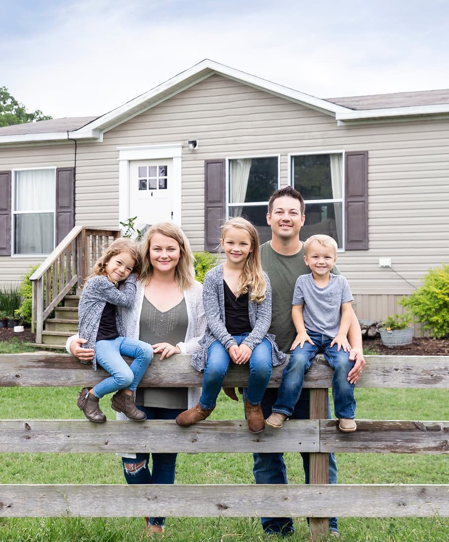 A happy homeowning family of five sit outside of their modern manufactured home on a fence.