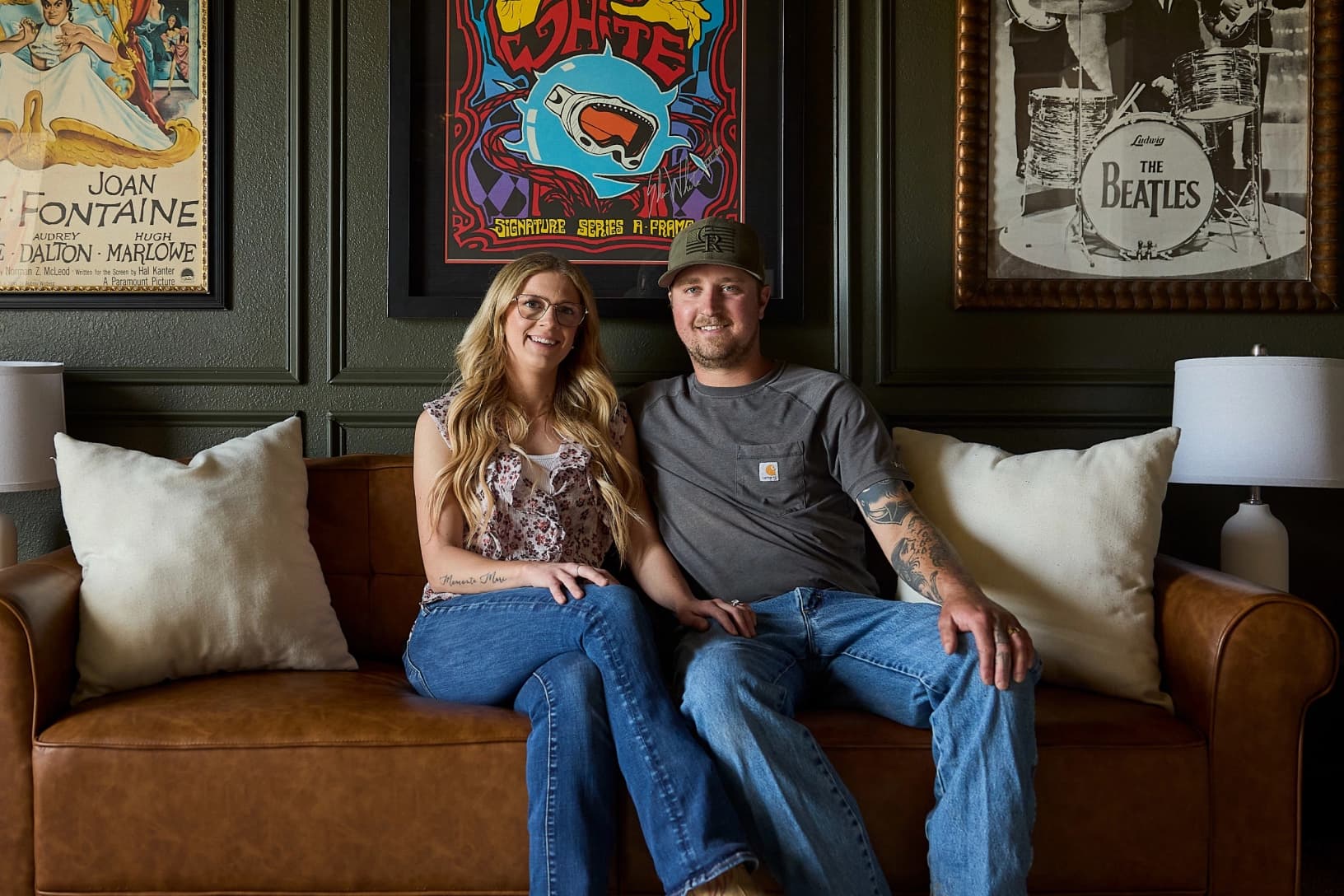 A couple sitting together on a leather sofa in a living room decorated with vintage posters.
