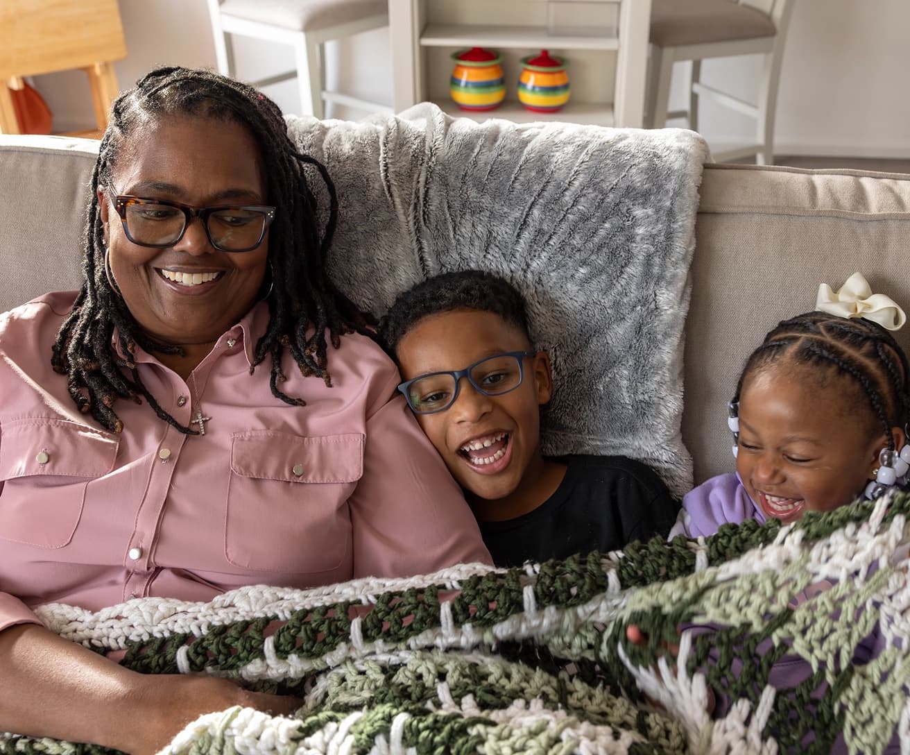 A smiling grandmother and two grandchildren laughing together under a blanket on the couch.