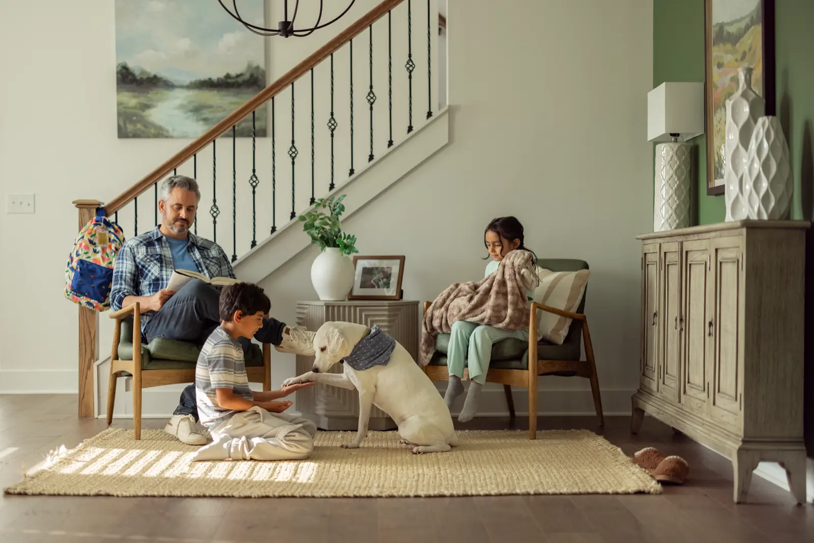 A family relaxing in their home's entryway, with a father reading, a boy petting the dog and a girl wrapped in a blanket.